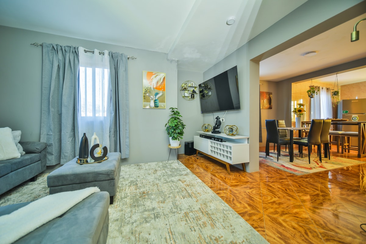 A comfortable living area is presented, featuring a plush gray sectional sofa and a large area rug. A flat-screen television is mounted on the wall, while natural light filters through a window with light gray curtains. A dining space is visible in the background.