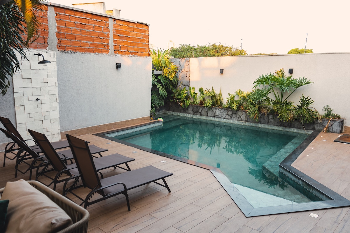 A large solar-heated pool is surrounded by a smooth deck. Several loungers are positioned nearby, offering a relaxing space for guests. Lush greenery surrounds the pool area, enhancing the tranquil setting and providing privacy from the neighboring spaces.
