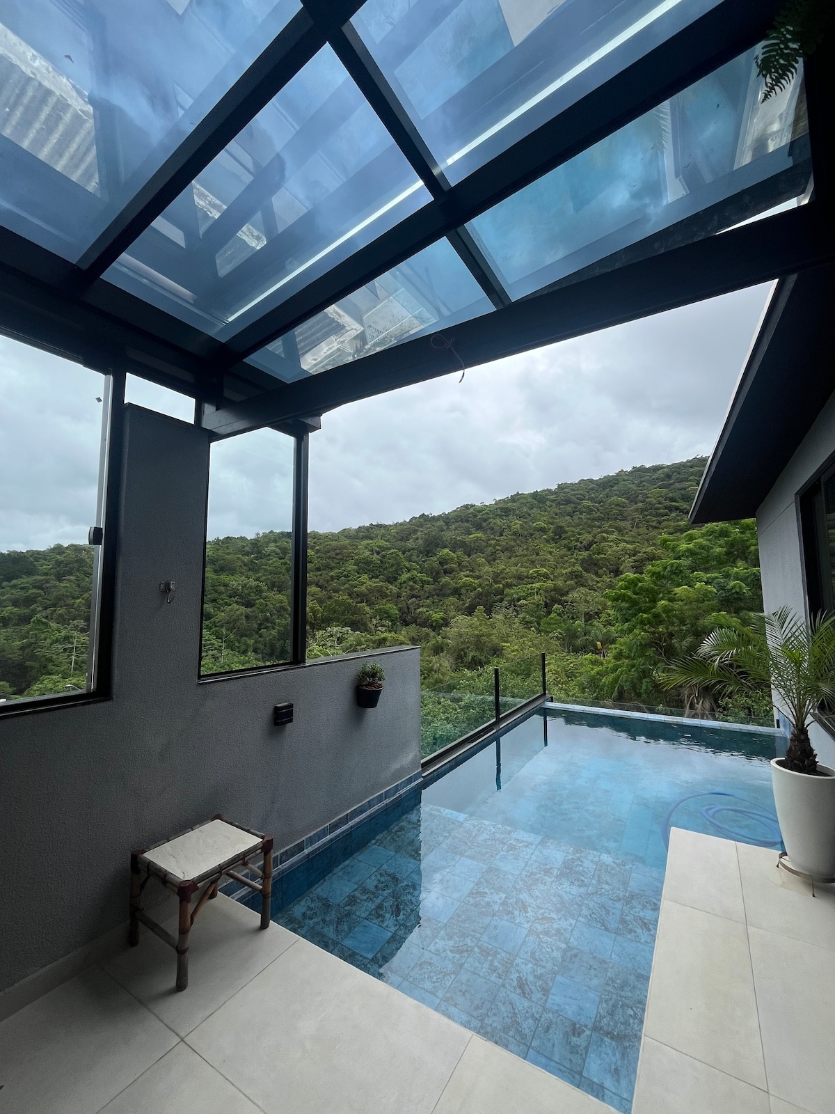 A serene outdoor space features a clear swimming pool bordered by a glass enclosure. Lush greenery is visible in the background, and a small wooden chair is placed nearby, offering a quiet spot to enjoy the view. Natural light filters through the blue-tinted roof.
