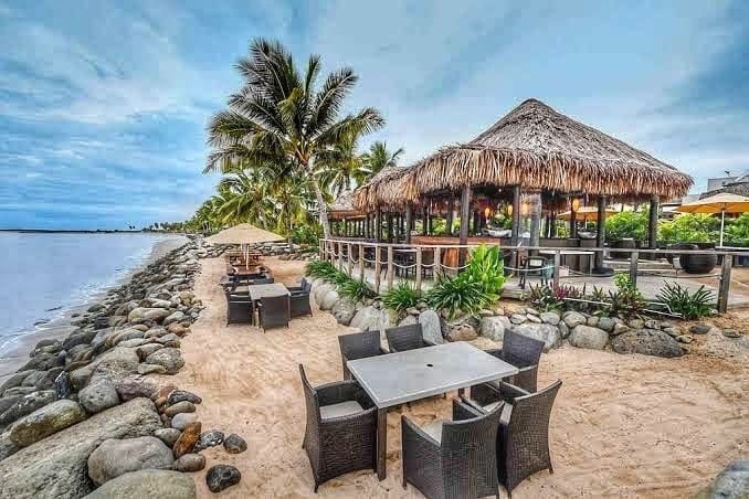 A beachfront restaurant features a thatched roof and large windows, surrounded by tropical palm trees. Outdoor seating includes tables and chairs on a sandy area, with decorative rocks lining the shoreline. An umbrella provides shade for guests, creating a relaxed dining environment.