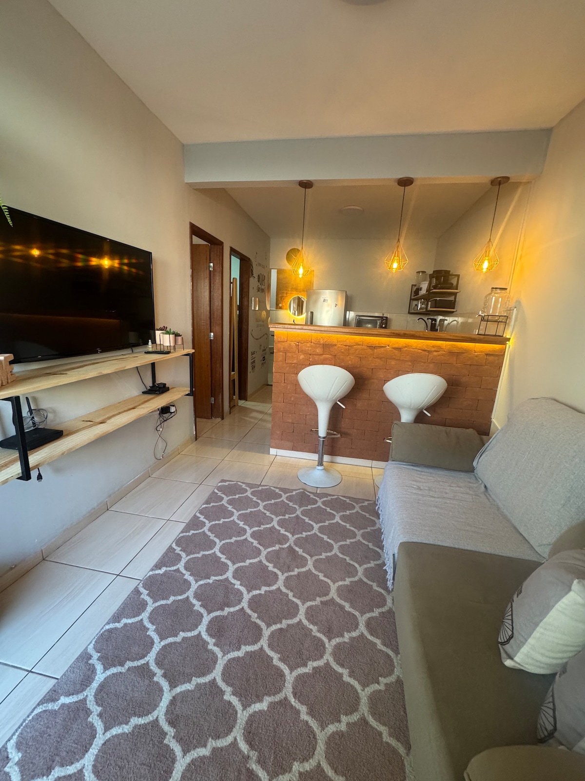 A modern living space features a neutral color palette with a comfortable sofa and a stylish rug. Two white bar stools are positioned at a small kitchen counter illuminated by pendant lights, and a flat-screen TV is mounted on the wall.