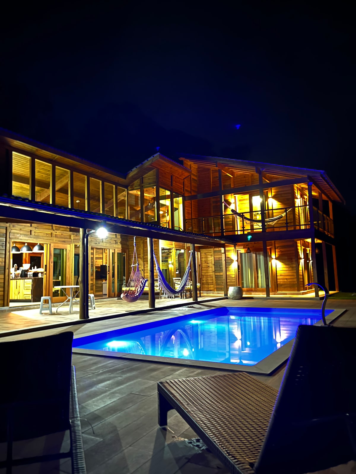 The exterior of a wooden house is illuminated against the night sky, highlighting a private pool surrounded by a deck. Lounge chairs are positioned nearby, and hammocks are visible hanging from beams. Large windows suggest a cozy indoor space filled with light.