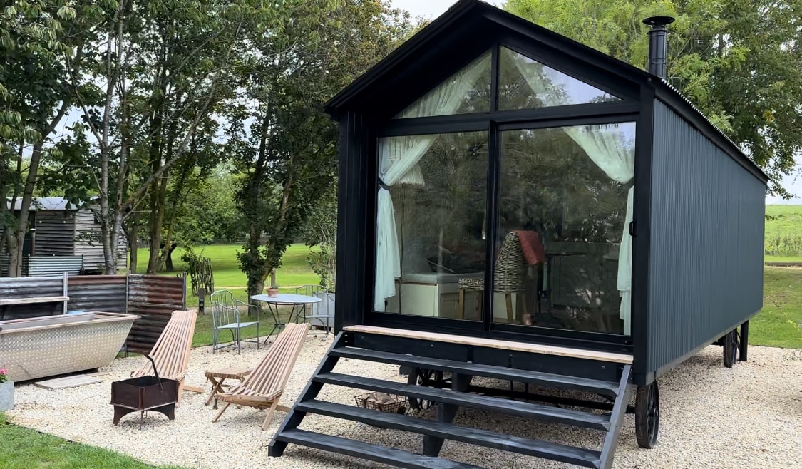 A stylish cabin with a sleek black exterior features large glass doors that offer views of the interior. Two reclined lounge chairs are placed on a pebbled patio, along with a small fire pit. Lush greenery surrounds the cabin, enhancing its natural setting.