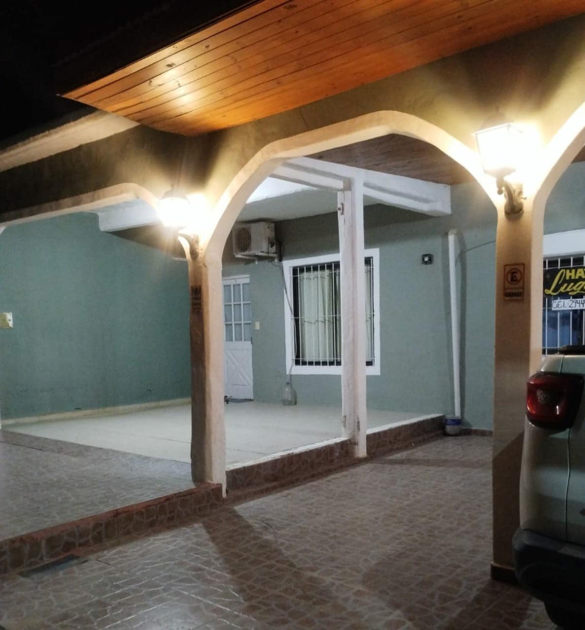 An entrance area features a spacious covered porch with sturdy columns and soft lighting provided by wall-mounted fixtures. The light-colored stone floor extends toward a well-defined entry point, while a nearby window is secured with bars for safety.