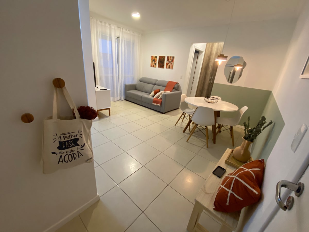 A welcoming open space is seen, featuring a gray sofa with orange and white accent pillows. A circular dining table with white chairs is positioned nearby. Natural light filters through sheer curtains, enhancing the bright and airy environment. Decorative items add subtle warmth to the space.
