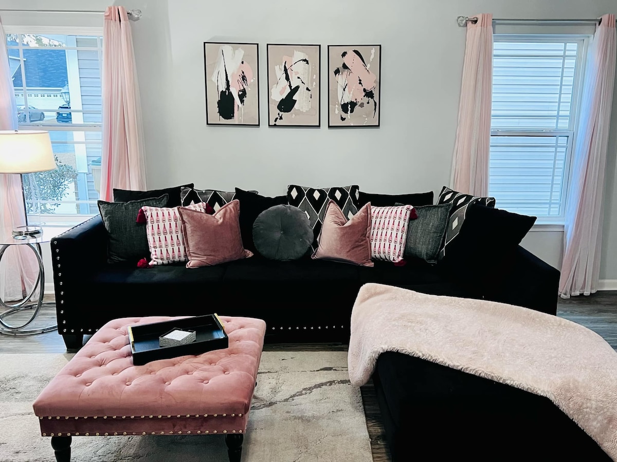 A living area features a black sectional sofa adorned with various cushions in shades of pink and gray. Above the sofa, three framed abstract artworks in black and pink are displayed. A soft pink ottoman and a light throw blanket add warmth to the space, complemented by pink drapes by the windows.