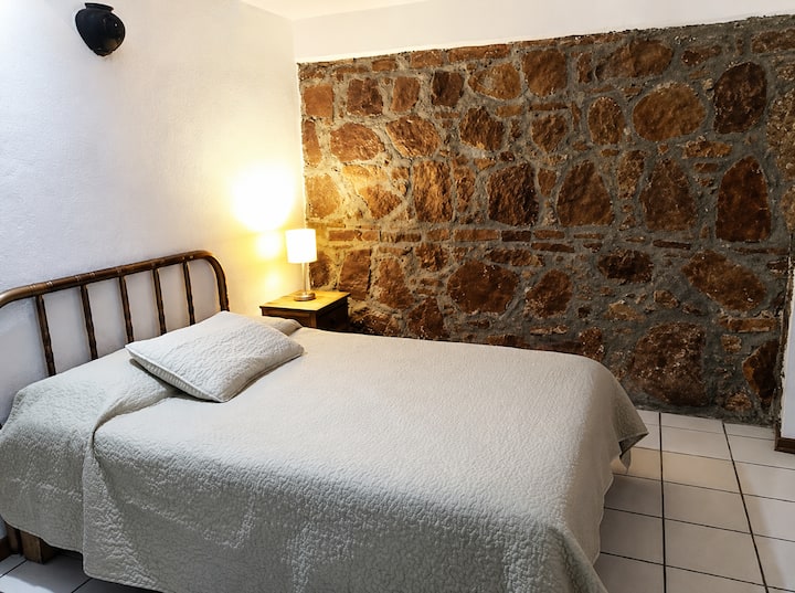 Mysigt sovrum med en autentisk kolonial stenmur, en charmig arkitektonisk detalj som är unik för RO-House. Dubbelsäng med nybäddade lakan och varm belysning för en perfekt vila i Oaxaca.