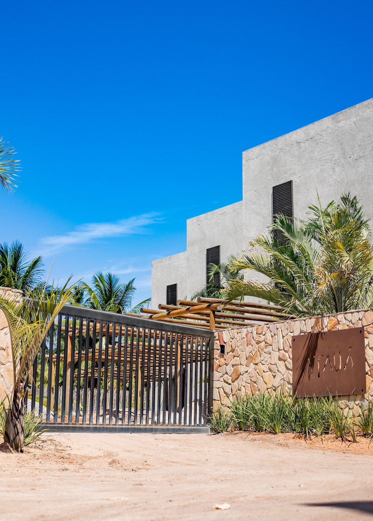 The entrance to the property is framed by a sleek metal gate and a natural stone wall. Lush greenery and palm trees add to the inviting landscape under a clear blue sky, setting the tone for a serene retreat.
