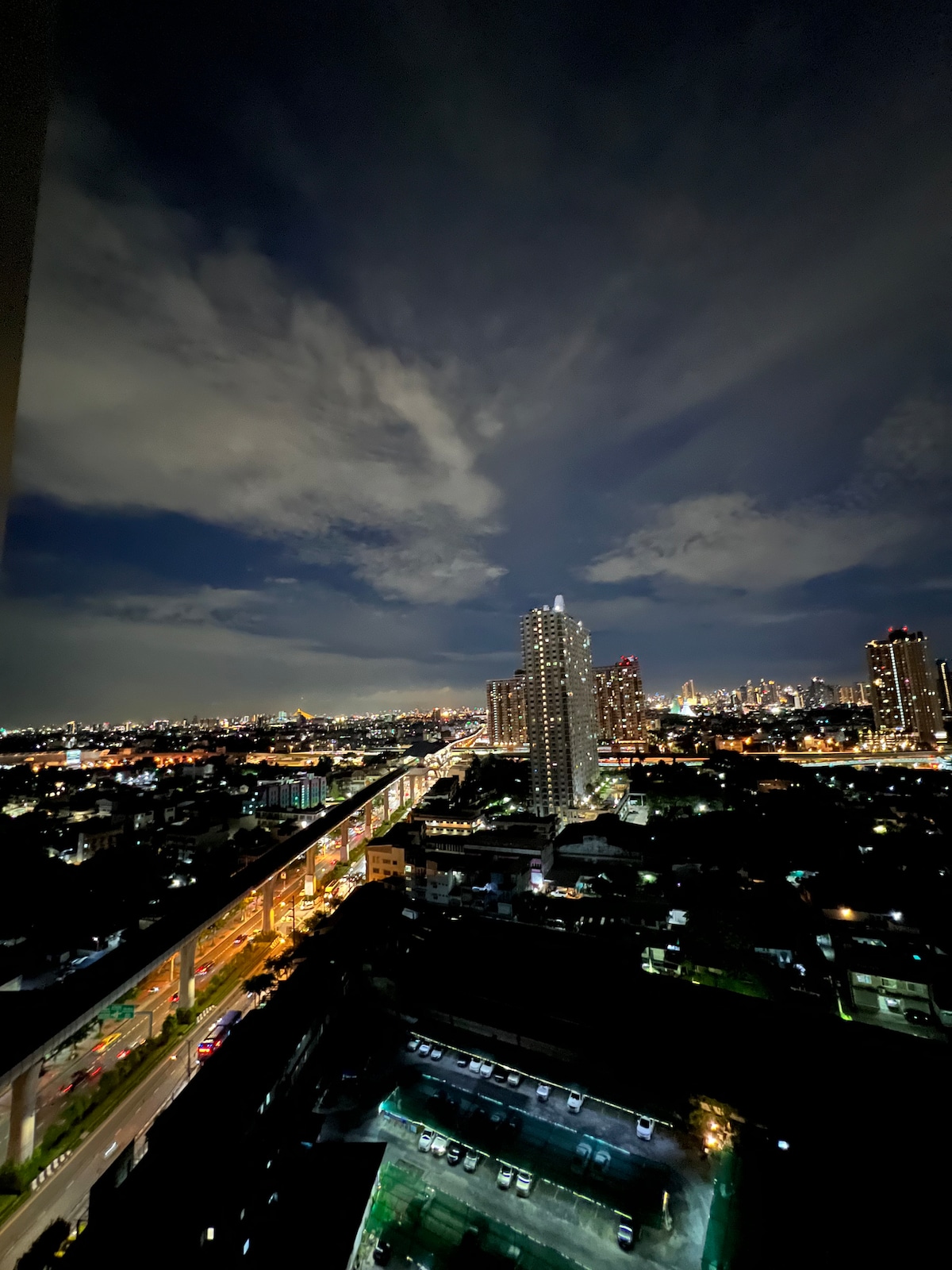A night view of a vibrant city skyline is displayed, featuring illuminated buildings and the winding path of a busy road below. Soft clouds drift across the night sky, creating a dynamic backdrop to the urban landscape, with city lights twinkling in the distance.