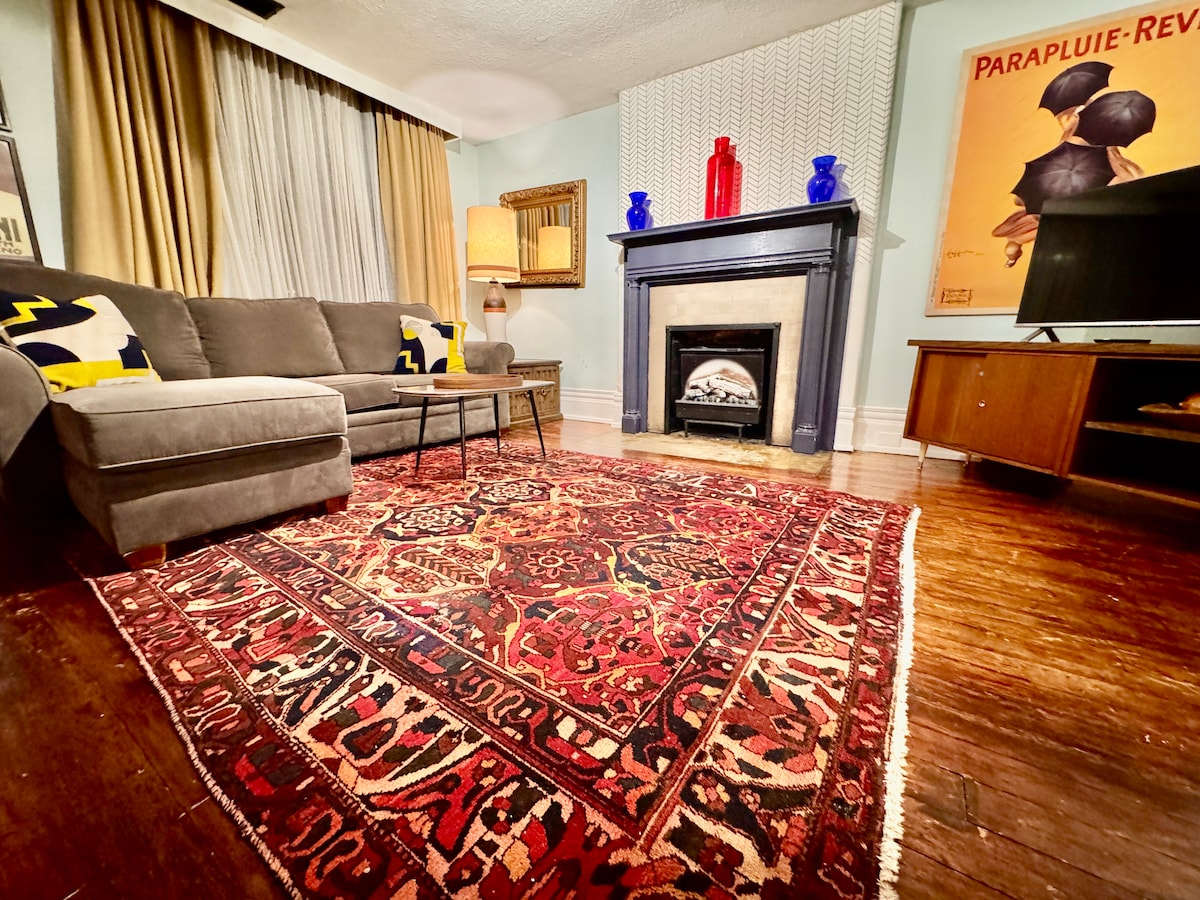 A comfortable seating area is highlighted by a plush gray couch and two decorative chairs, arranged around a classic fireplace. The hardwood floor is adorned with a rich, colorful area rug, while light filters in through soft drapes, enhancing the room's inviting ambiance.