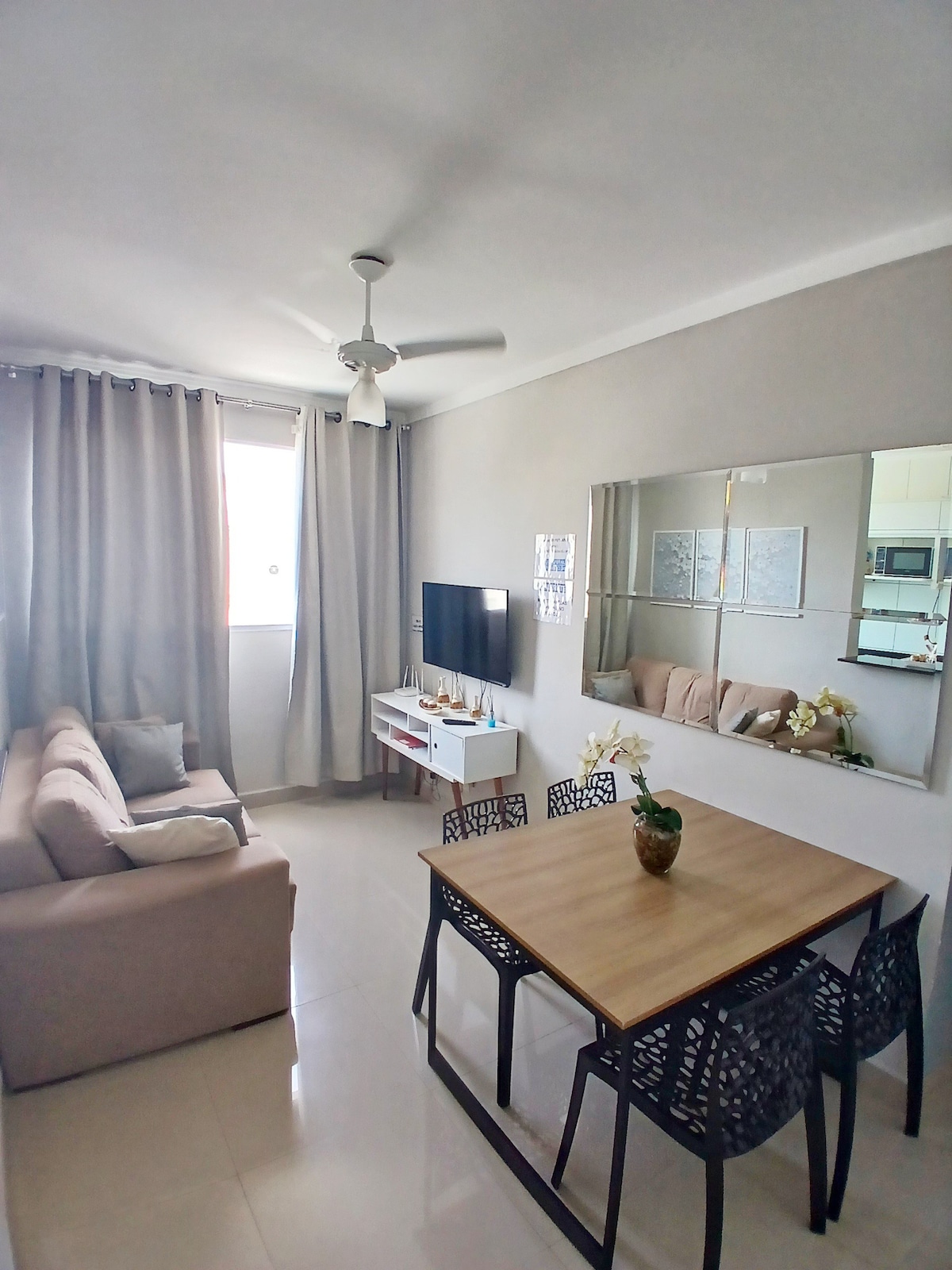 A modern living area features a comfortable light-colored sofa and a dining table with black chairs. A wall-mounted television is positioned across from the seating, and natural light enters through large windows covered by light curtains. A mirror enhances the space's brightness.