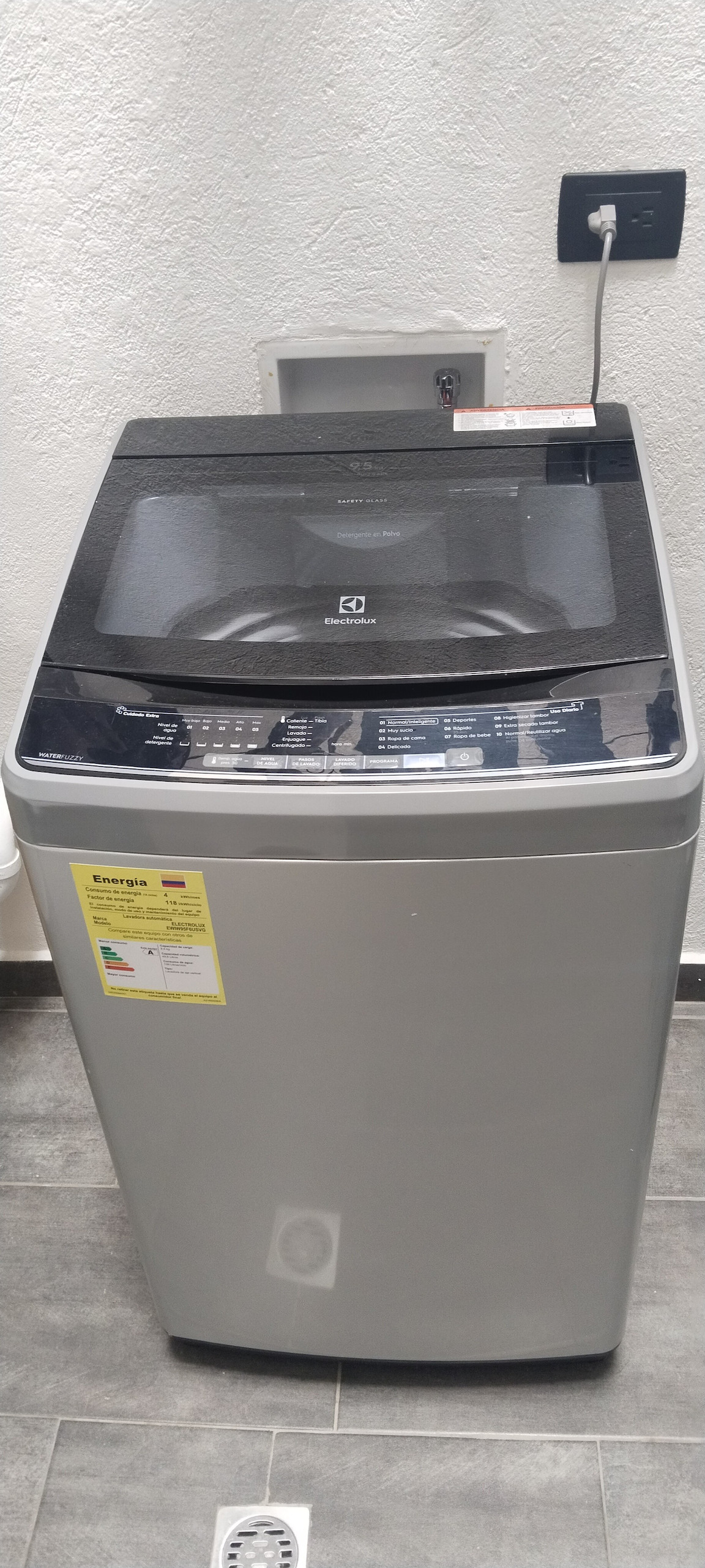 A modern washing machine is positioned against a textured wall. The top-loading machine features a sleek silver exterior and a digital control panel at the top. A yellow energy efficiency label is attached, indicating compliance with energy standards.