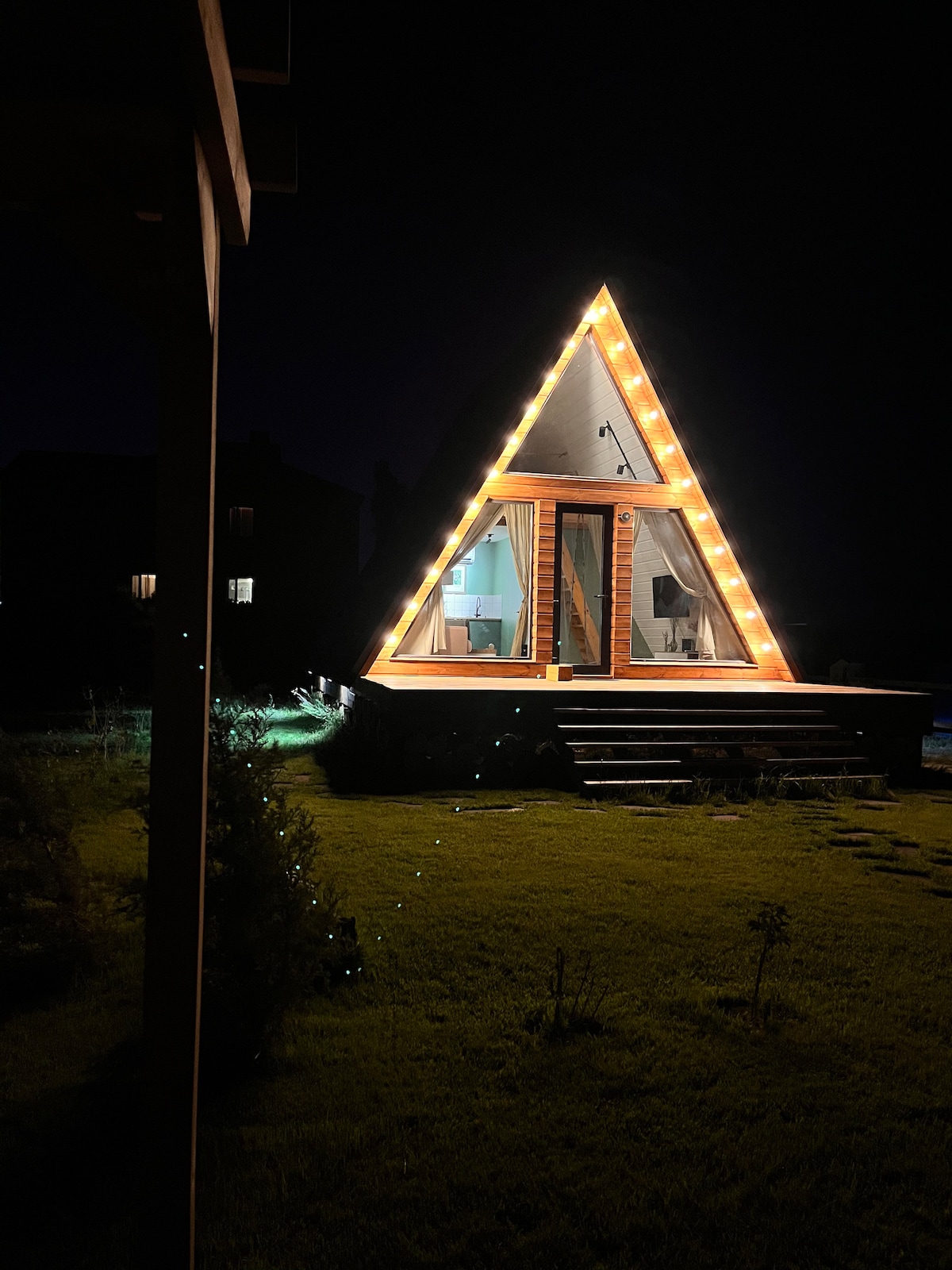 The A-frame cabin is illuminated by warm outdoor lighting, highlighting its unique triangular shape against the night sky. Soft curtains frame large glass doors, which provide an inviting entrance. The surrounding greenery is visible, offering a serene contrast to the cabin's bright exterior.
