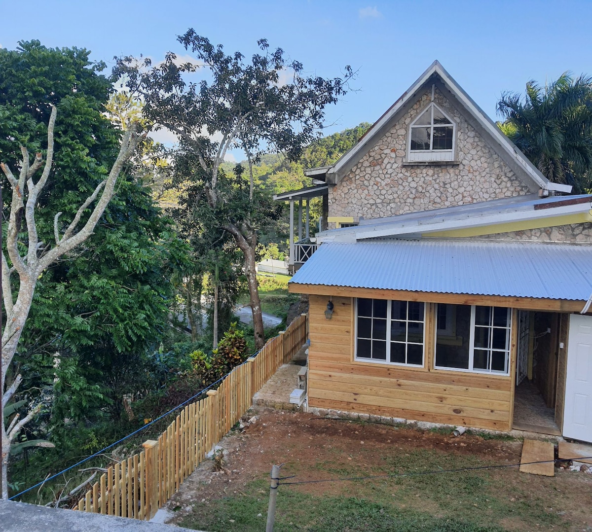 A charming stone and wooden structure is surrounded by lush greenery, with a fenced area and pathway visible. The house features a gabled roof and large windows, providing ample natural light, while the nearby trees add to the serene rural atmosphere.