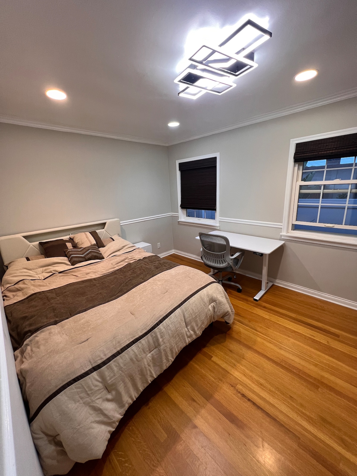 A well-lit bedroom features a spacious bed with neutral bedding and decorative pillows. A sleek desk is positioned next to the window, where natural light enters through partially shaded blinds. Warm wood flooring complements the room's overall design.