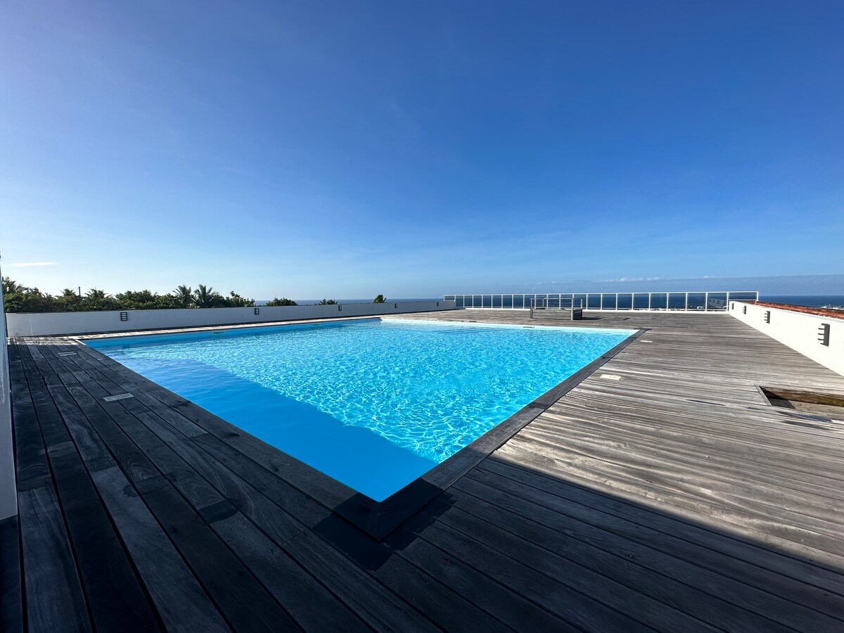 An expansive swimming pool is featured on a wooden deck, reflecting the bright blue sky. Surrounding greenery is visible in the distance, creating a serene outdoor atmosphere.