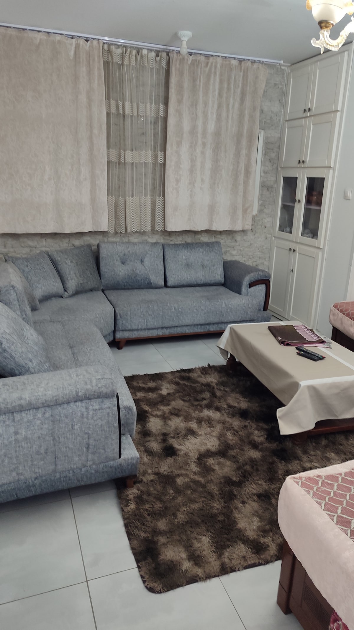 A comfortable living area is featured with a gray sectional sofa and a soft brown rug. A low coffee table sits in front, while sheer curtains filter light from the window. Nearby, a compact dining area is visible, adorned with a light-colored tablecloth.