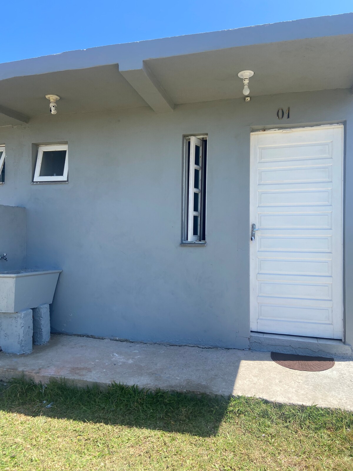 An exterior view presents a simple single-unit building with a light gray façade. The entrance is framed by a white door. Two small windows are positioned nearby, allowing natural light. A concrete area is noticed to the left, with a basic sink installed, and grass covers the ground.