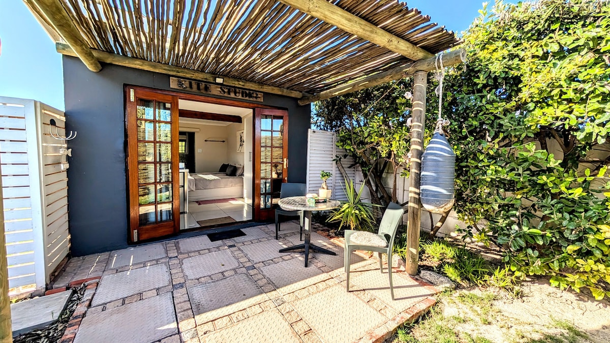 An outdoor veranda is shaded by a wooden overhang, with a small table and two chairs arranged on stone paving. Lush greenery surrounds the area, and a hammock hangs nearby, inviting relaxation in the tranquil setting.