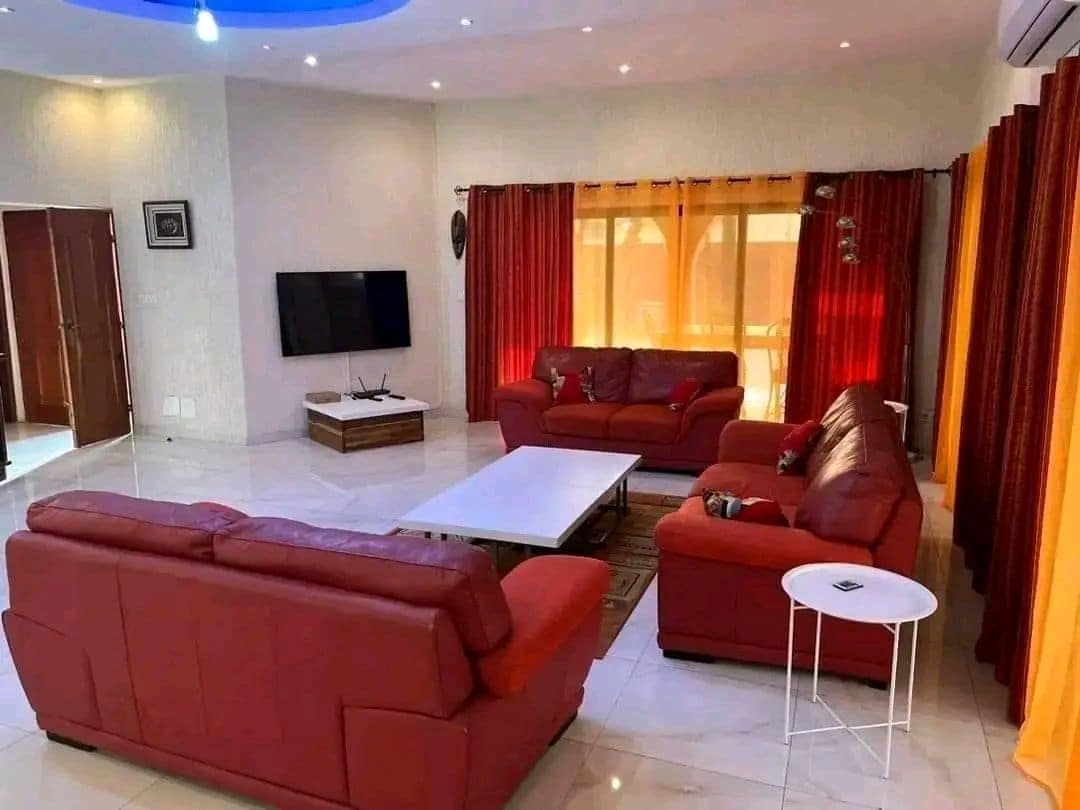 A bright and spacious living area features a large white coffee table surrounded by deep red leather sofas. Large windows are adorned with flowing curtains, allowing natural light to fill the room. A sleek television is mounted on the wall opposite the seating arrangement.