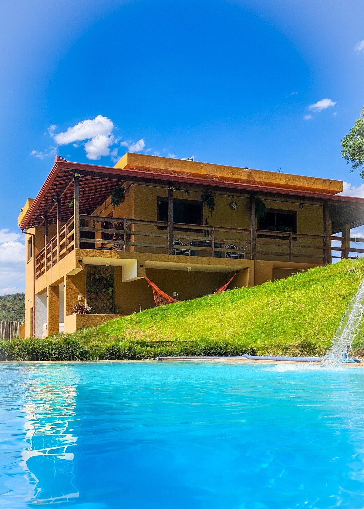 The exterior of the property is visible, showcasing a two-story building with a wooden balcony and large windows. A vibrant green lawn extends toward a pool, reflecting sunlight. The sky is clear, enhancing the inviting outdoor setting.