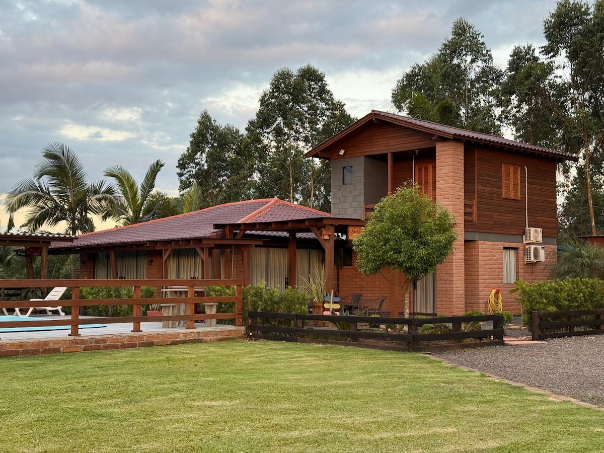A welcoming exterior of a spacious property is depicted, showcasing two distinct structures surrounded by lush greenery. An inviting swimming pool and lounging area are visible, with a wooden fence framing the features. The landscape is well-maintained, providing a serene atmosphere.