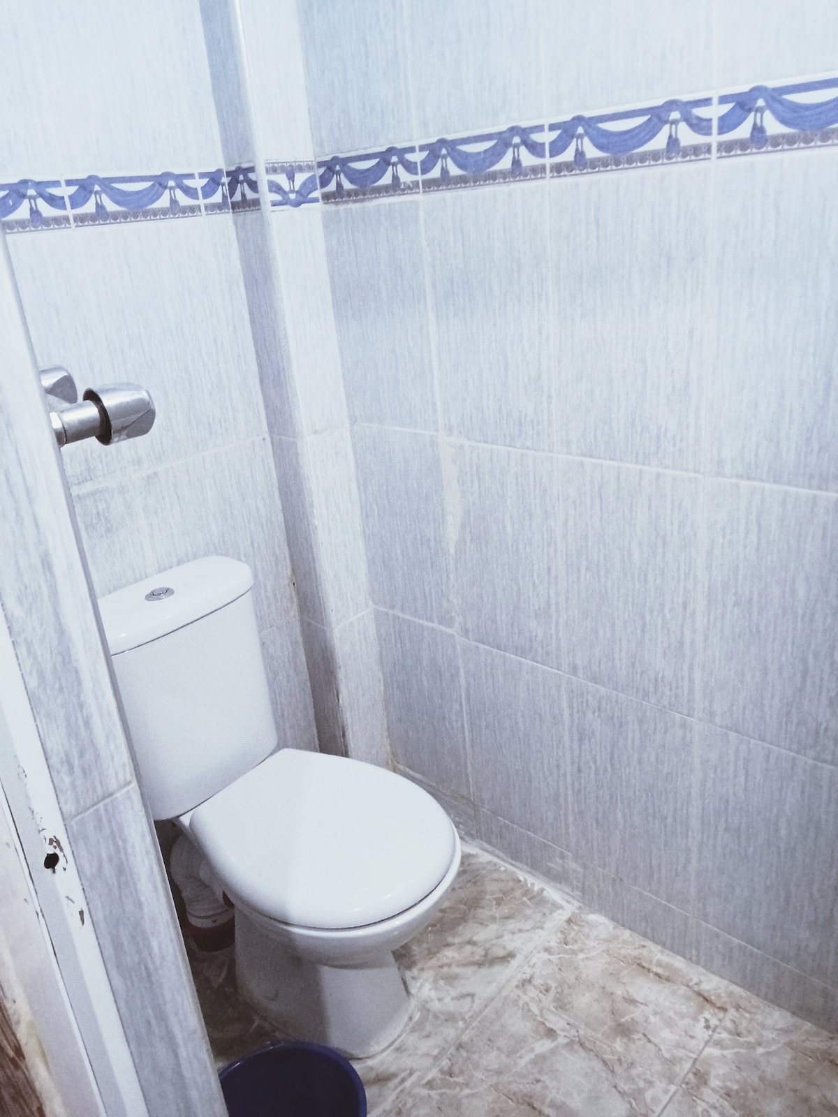 A compact bathroom features a white toilet against light grey tiled walls. A decorative border runs along the top, adding a subtle touch. The floor is covered in light-colored tiles, and a small bin is visible in the corner.