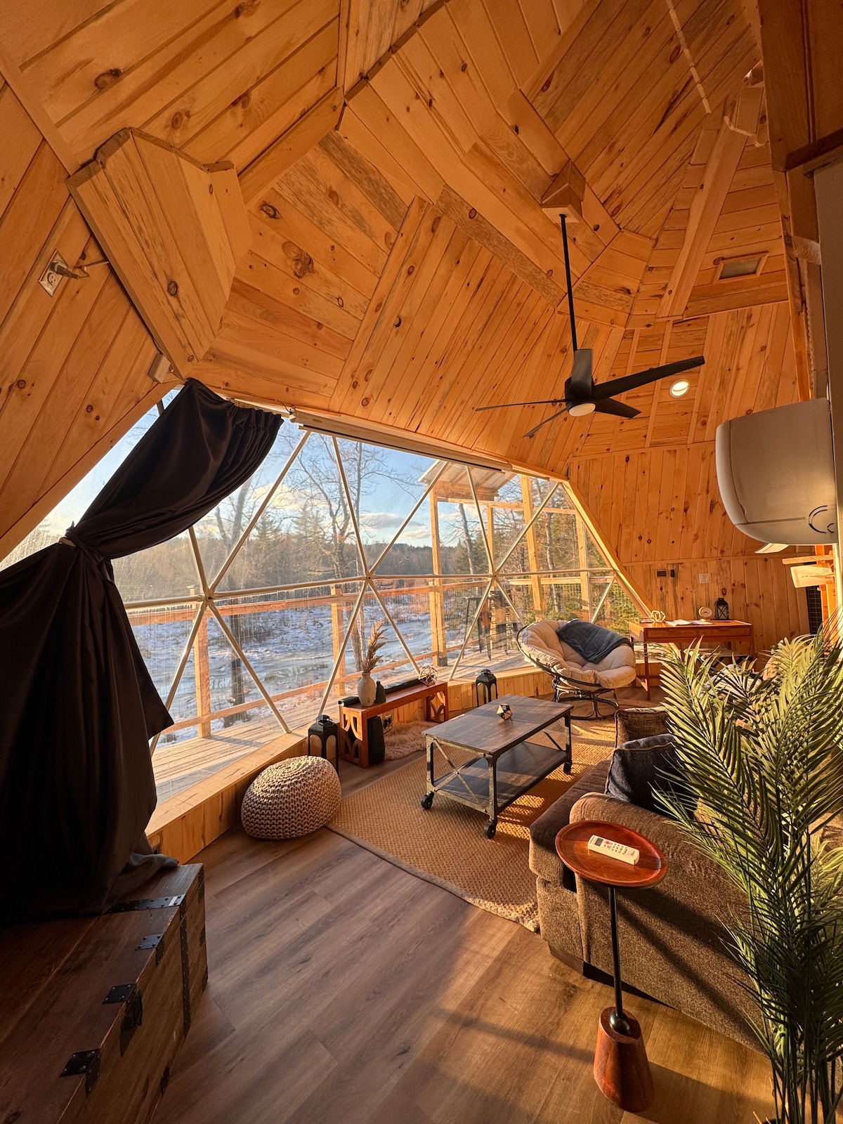An open living area features wooden walls and a geometric glass design. Comfortable seating is arranged around a low coffee table. Natural light floods the space, highlighting views of the surrounding landscape through large windows.