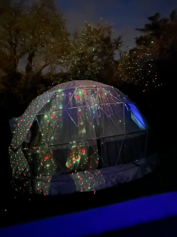 A geodesic dome is illuminated with soft, colorful lights against a darkened sky, creating a serene ambiance. The dome features transparent panels that showcase the playful light reflections, surrounded by trees, enhancing the peaceful outdoor setting.