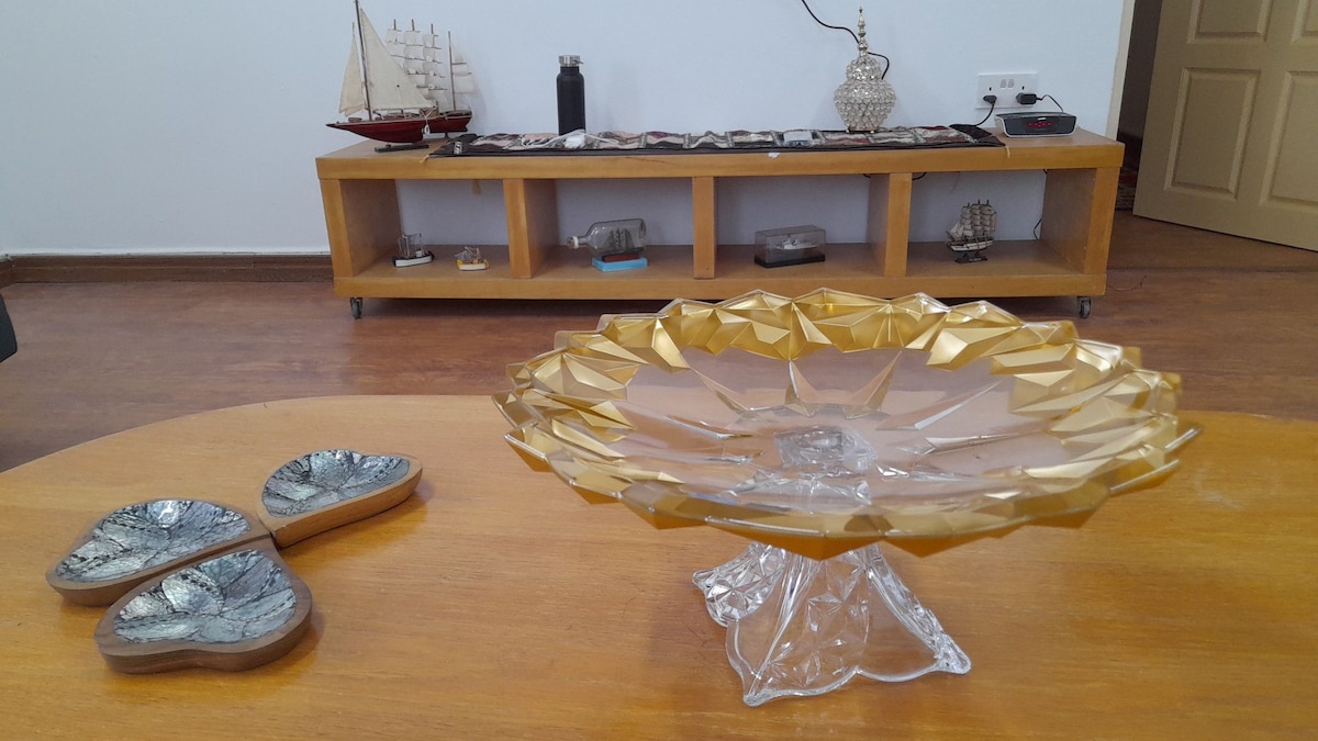 A decorative glass plate with a gold geometric design rests on a wooden table. Nearby, two leaf-shaped ceramic dishes are displayed. In the background, a wooden shelf holds various decorative items, including a model ship and a glass figurine.