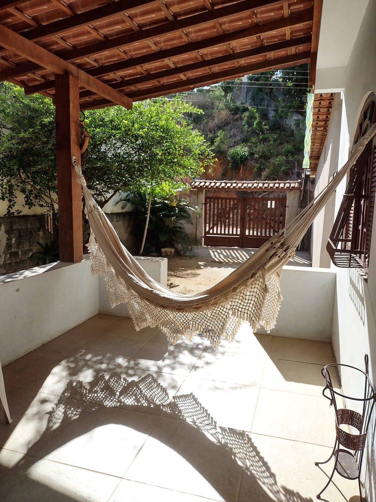 A relaxing outdoor space is featured, showcasing a hammock suspended between wooden posts. Light filtering through the trees casts gentle shadows on the tiled floor. A welcoming entrance with a wooden gate and lush greenery completes the serene atmosphere.