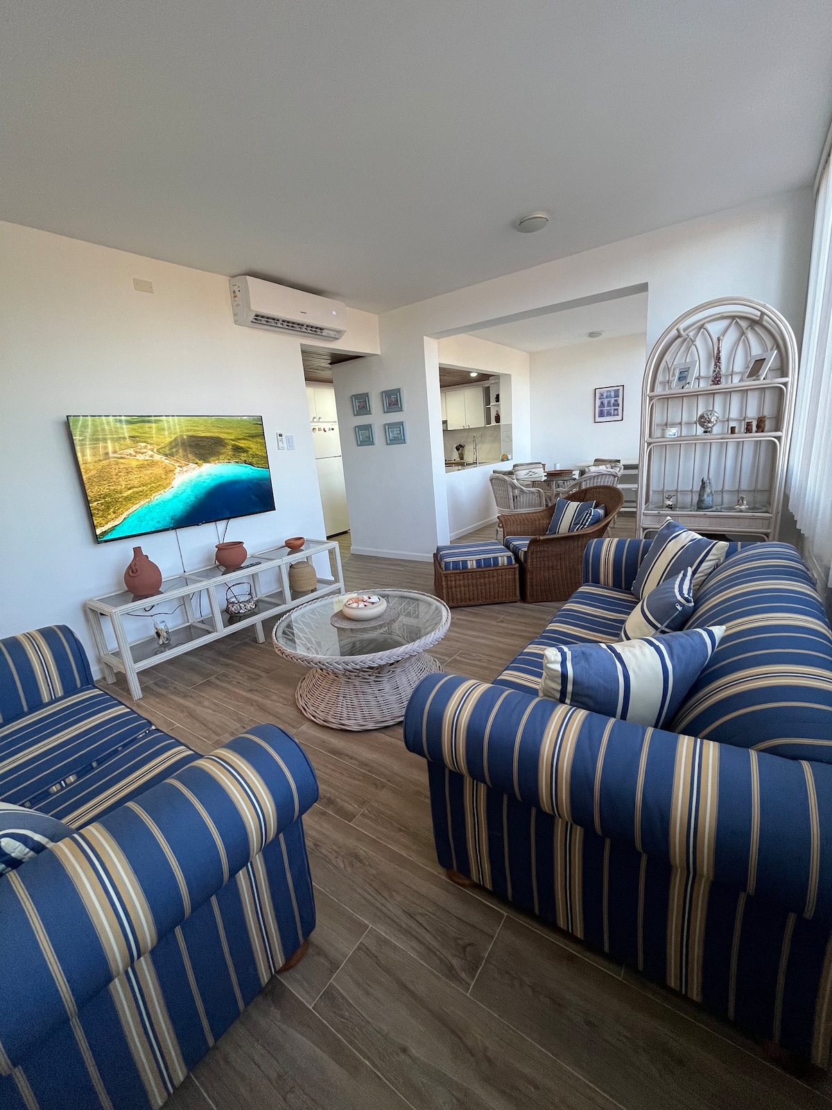 A comfortable living area is furnished with striped blue sofas, complemented by woven accent chairs. A circular coffee table sits in the center, adorned with a decorative item. A mounted television displays a nature scene, and natural light brightens the space through large windows.