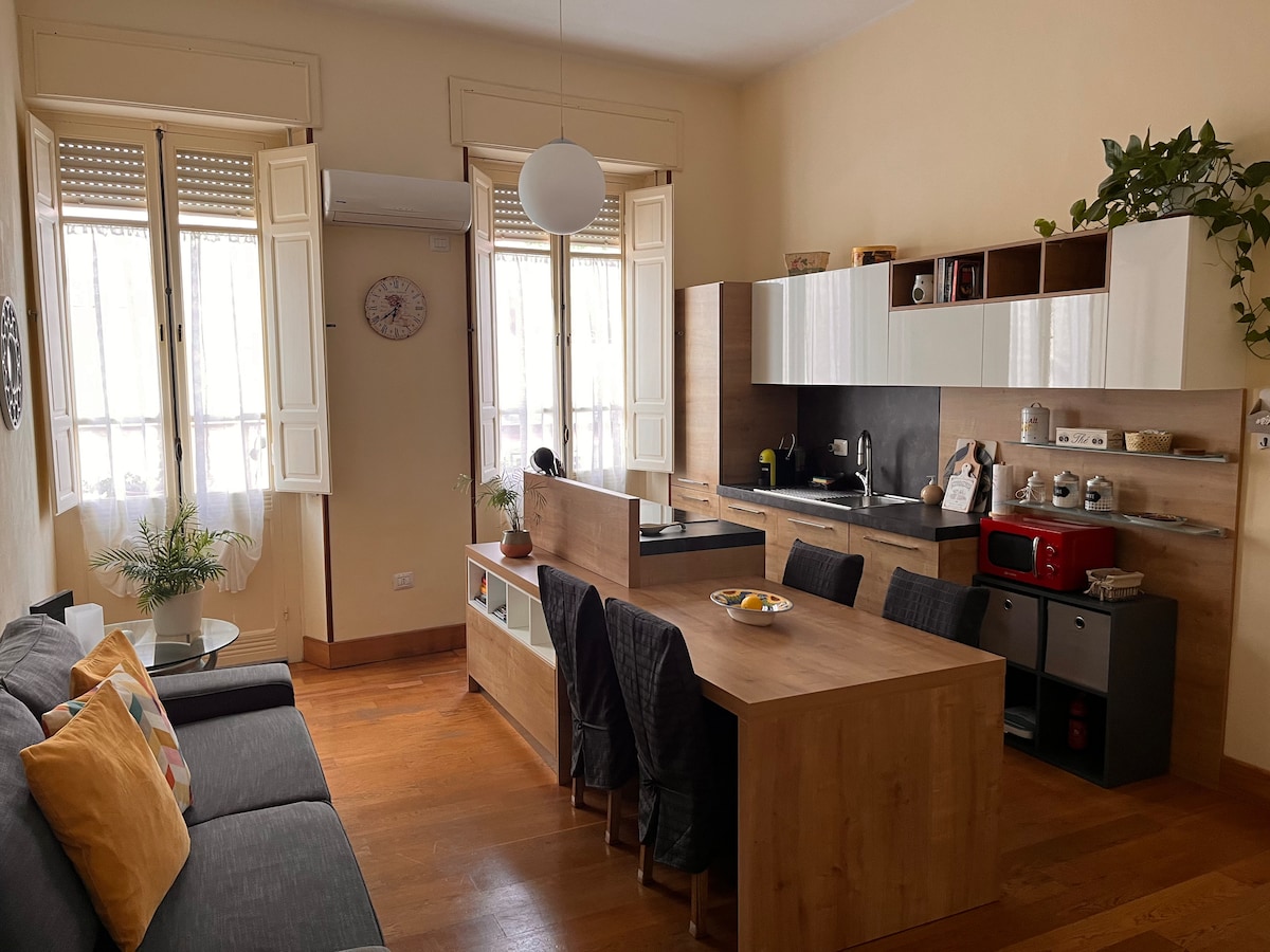 A modern kitchen area is visible, featuring a wooden dining table with black chairs. Open cabinets display kitchenware and a red microwave. Two large windows provide natural light, complemented by light-colored walls. A cozy seating area with a dark sofa and colorful cushions is also present.