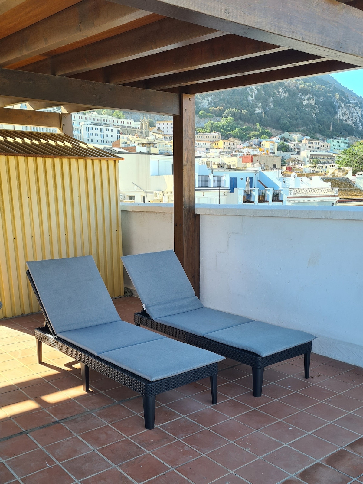 Two lounge chairs are positioned on a tiled rooftop terrace, offering a partial view of the surrounding buildings and hillside. The area is shaded by a wooden pergola, with a storage shed visible in the background.