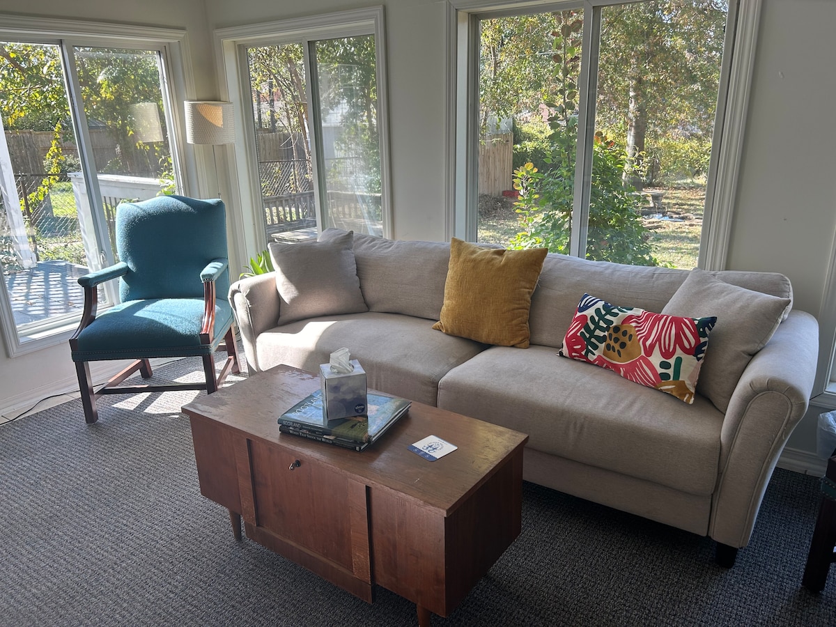 A sunlit sun room features a comfortable sectional sofa accompanied by a colorful throw pillow. A wooden coffee table sits in front, with a small decorative box on top. Windows allow natural light to filter in, offering views of the outdoor greenery.