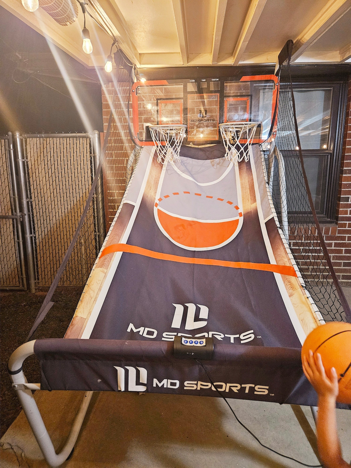 An indoor basketball game is positioned under a cover, featuring two hoops at opposite ends of a long playing surface. A digital scoreboard is mounted at the bottom, while soft lighting from overhead fixtures enhances the playful atmosphere.