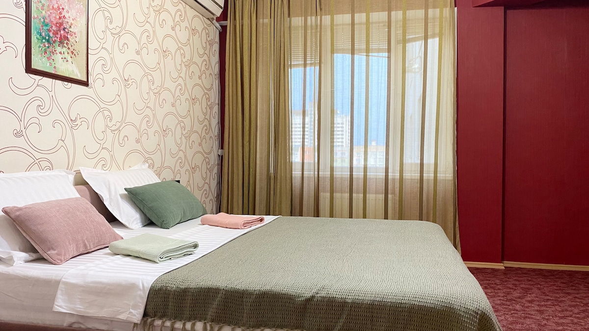 A spacious bedroom featuring a large bed with light linens and a decorative green throw. Two sets of pillows in soft colors are placed neatly. Sheer curtains allow natural light to filter in, complementing the warm tones of the wall decor.