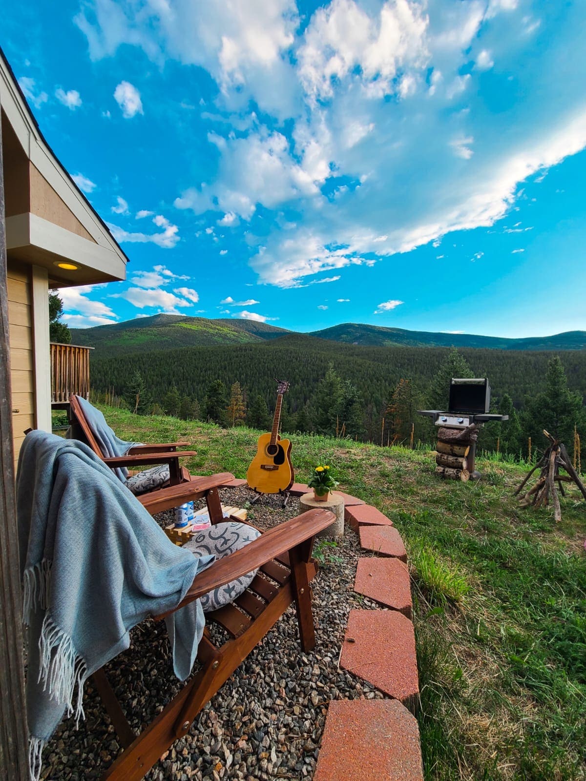 A serene outdoor space features two wooden chairs adorned with soft blankets, set against a backdrop of expansive mountain views. A guitar rests nearby, while a grill is positioned a short distance away. Lush green foliage complements the vibrant blue sky.