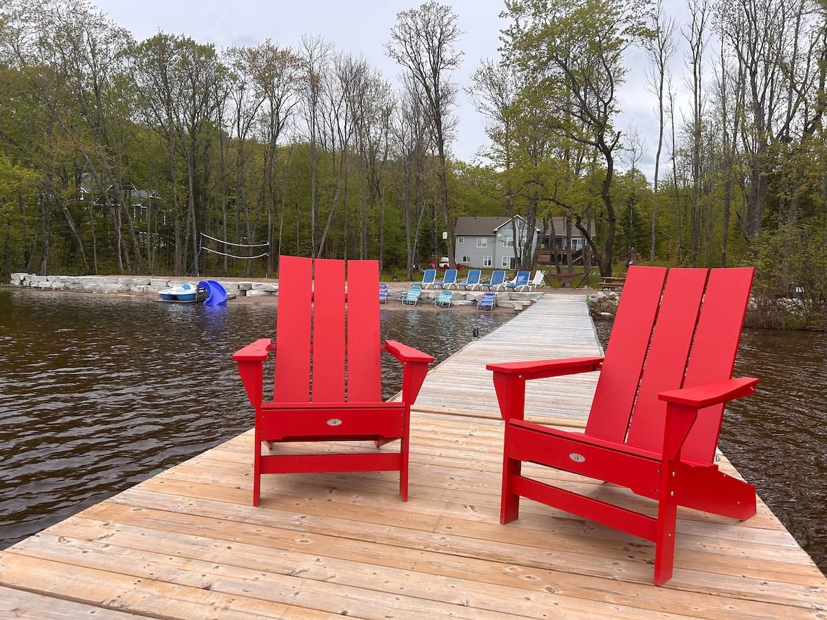 Two bright red Muskoka chairs are positioned on a wooden dock overlooking a calm lake. In the background, various watercraft can be seen alongside a row of lounge chairs, with trees lining the shore in a natural setting.
