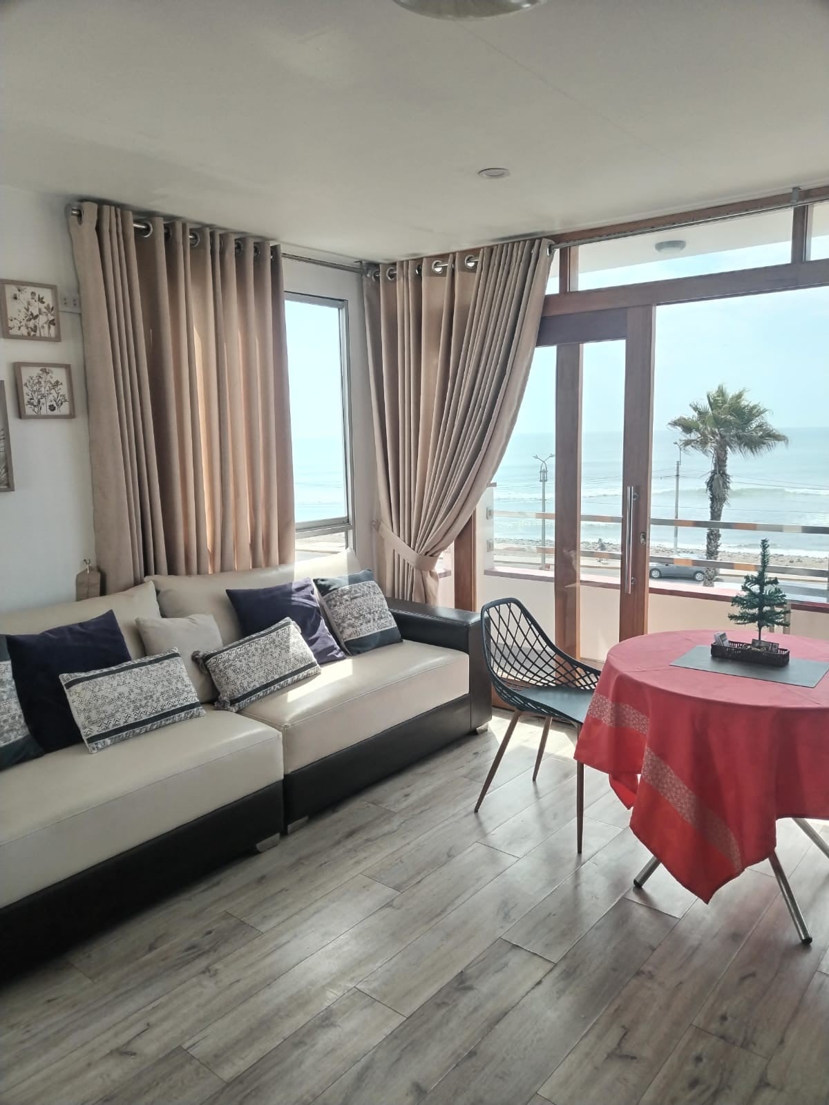 A spacious living area features a comfortable sofa with decorative pillows and a round table set for dining. Large windows provide abundant natural light and offer a view of the ocean and a palm tree swaying outside. Light-colored curtains add softness to the space.