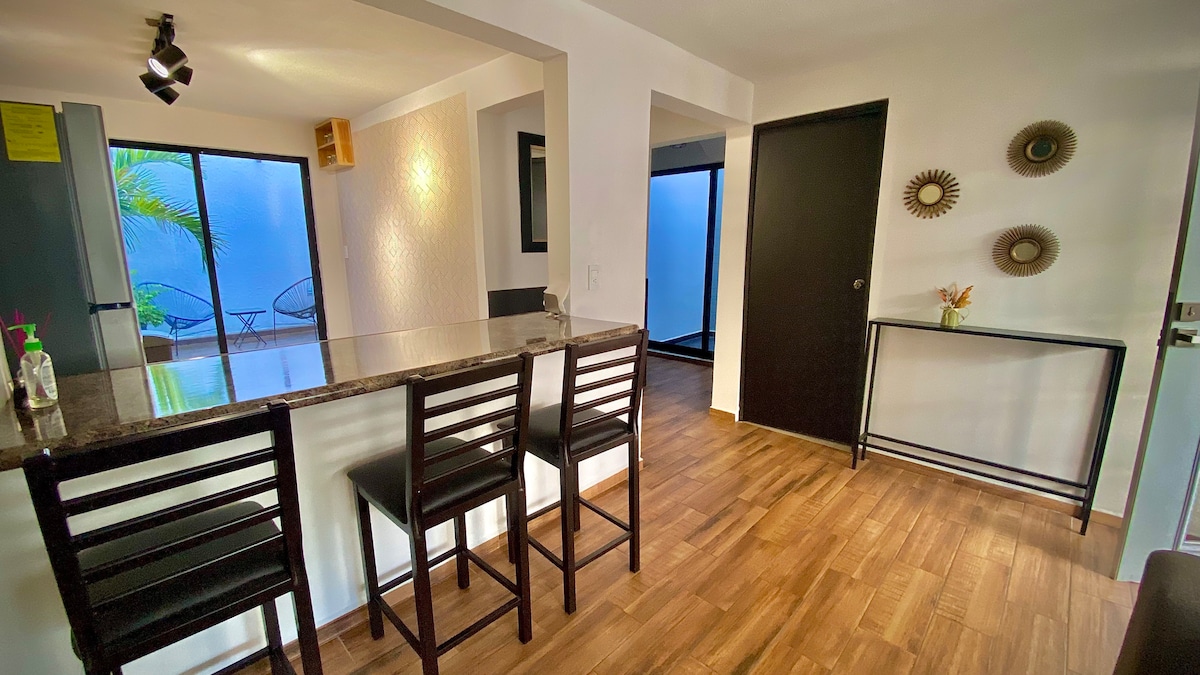 A modern kitchen area features a granite countertop with four wooden stools arranged along the side. Natural light enters from a large glass door leading to an outdoor space, complemented by light-colored walls and warm wood flooring throughout the area.