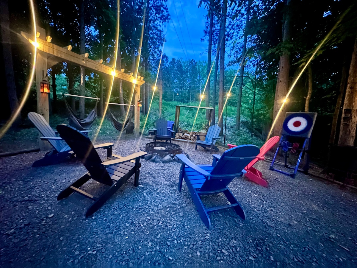 A fire pit area is surrounded by colorful Adirondack chairs in a wooded setting. String lights are draped overhead, providing a warm glow in the evening. Nearby, a hammock and a target stand add to the outdoor ambiance, with tall trees framing the space.