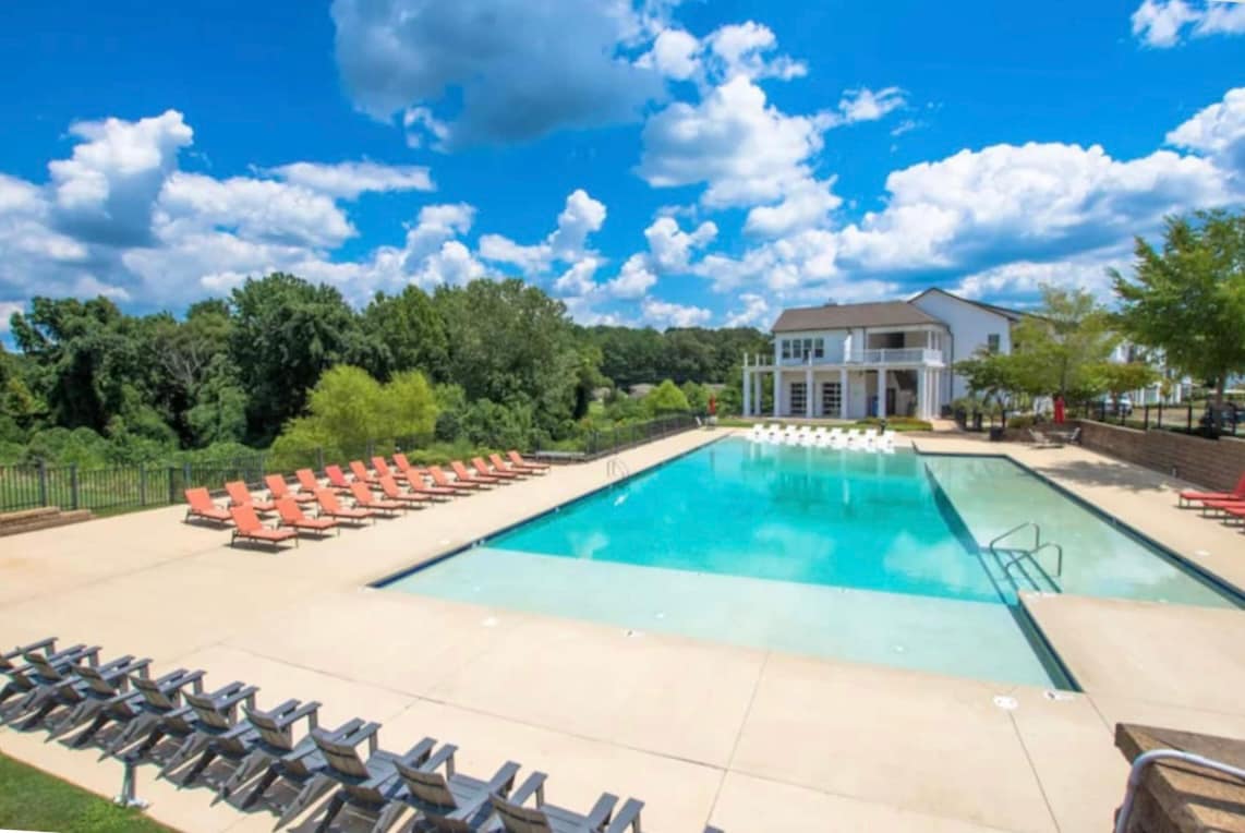 An expansive outdoor pool area is surrounded by lounge chairs, arranged in orderly rows. A clubhouse with large windows overlooks the pool, while lush greenery lines the background. The sky is clear, dotted with fluffy clouds, creating a bright and inviting atmosphere.