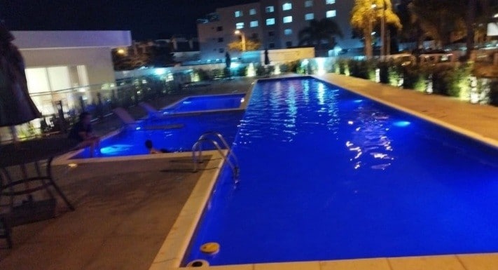 A well-lit pool area showcases two swimming pools under a night sky. The water reflects bright blue hues, while lounge chairs line the edge. Surrounding greenery creates a serene atmosphere, complemented by soft illumination from the nearby buildings.