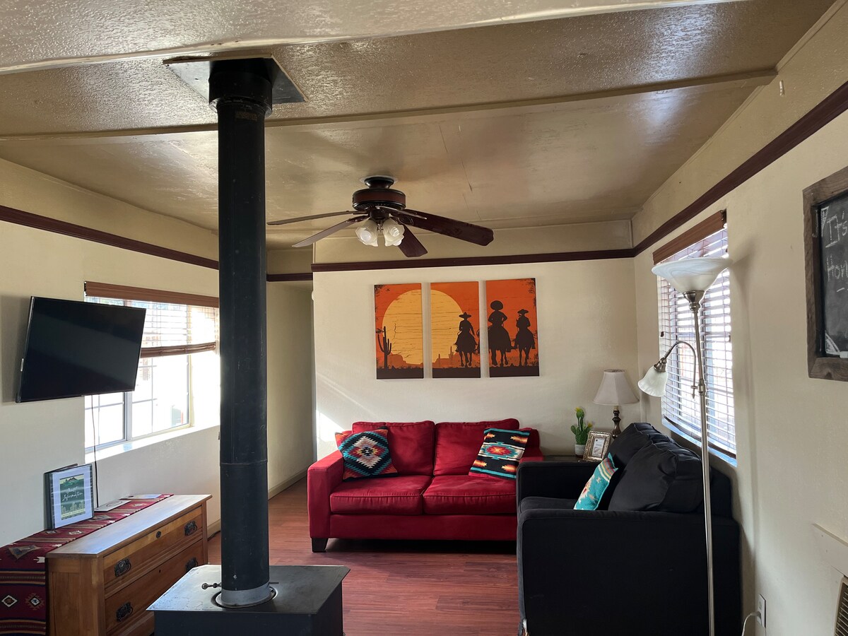 A cozy living area is equipped with a red sofa and a dark chair. A large artwork featuring silhouettes against a sunset is displayed above. A ceiling fan and wall-mounted television are present, while a wooden chest and lamp add to the inviting space.