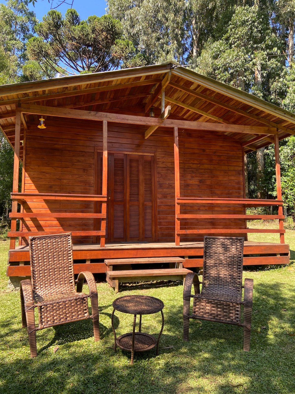 A cabin constructed from wood is set in a green landscape. The structure features a sloped roof and a wooden porch with steps leading to the entrance. Two woven chairs and a small table are placed on the grass in front, offering a cozy seating area.