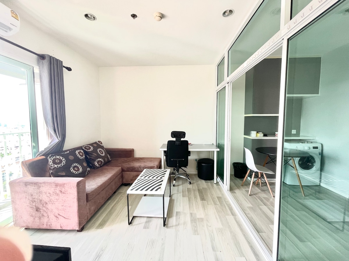 A modern living area features a plush sectional sofa adorned with patterned cushions. A sleek desk and ergonomic chair are positioned beside large glass windows that allow natural light. A minimalistic coffee table sits in front of the sofa, enhancing the space's open feel.