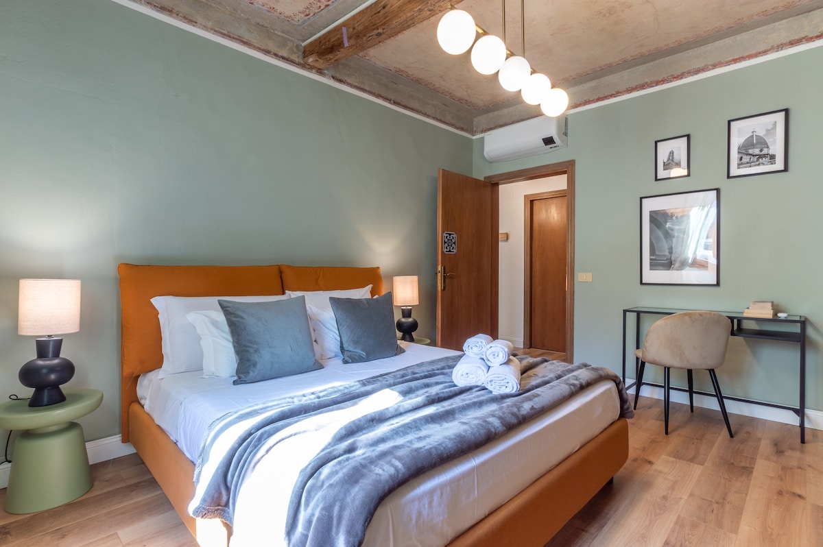 The bedroom features a spacious bed with soft pillows and a cozy grey throw. Two bedside lamps provide warm lighting, while framed art hangs on the wall. A small desk with a chair complements the room, which showcases a muted green wall and wooden ceiling beams.
