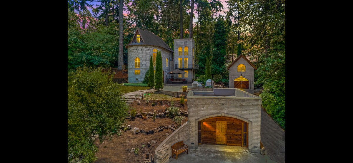 The exterior of a castle-like structure is framed by lush greenery, highlighted by warm outdoor lighting. A turret rises prominently, accompanied by multiple windows reflecting the soft glow of evening. A well-maintained landscape leads to a stone carriage house, enhancing the property's unique charm.