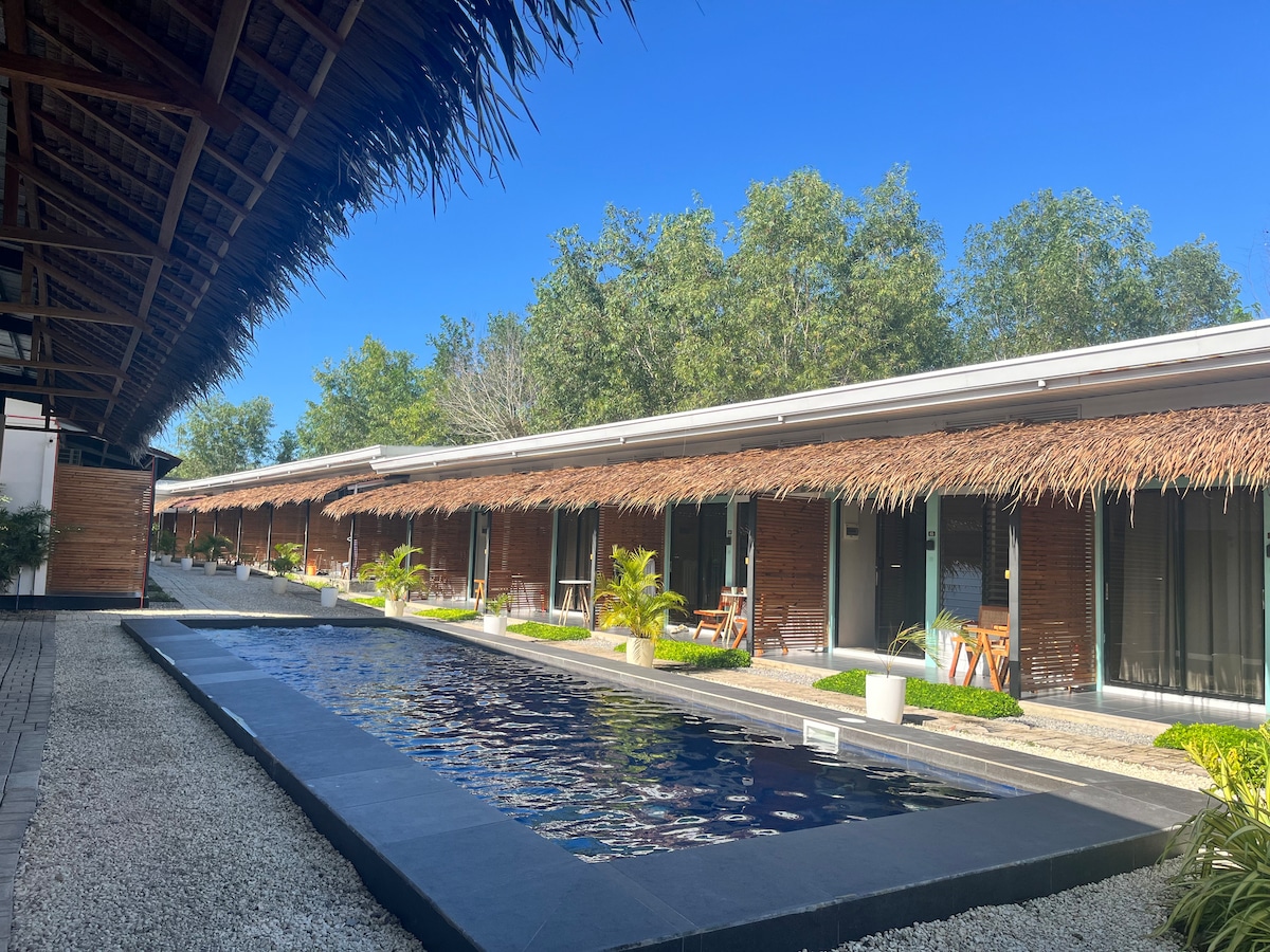 A tranquil courtyard is framed by charming thatched-roof structures, featuring access to a narrow swimming pool. Lush greenery surrounds the pool area, enhancing the peaceful ambiance of the space. Sunlight filters through the trees, creating a serene atmosphere.